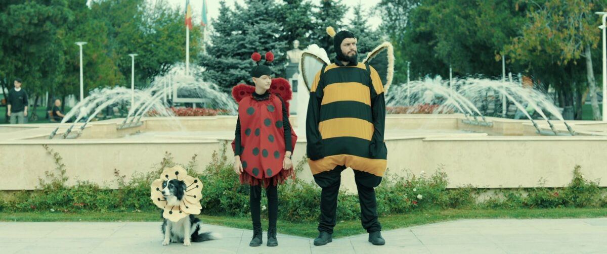 Two actors in ladybird and bee costumes, next to a dog in costume, in a park with fountains