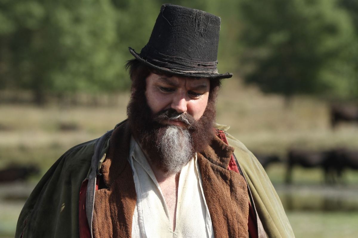 A portrait of a character from the film, a man with a tall hat and a beard, against a field background